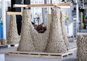 La imagen ilustra un robot depositando materiales como arcilla y tierra excavada para construir una estructura sin usar cemento.